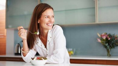 smiling happy woman eating food in kitchen 390x220 - چگونه کمتر غذا بخوریم؟ 15 راه مهار گرسنگی و کاهش اشتها